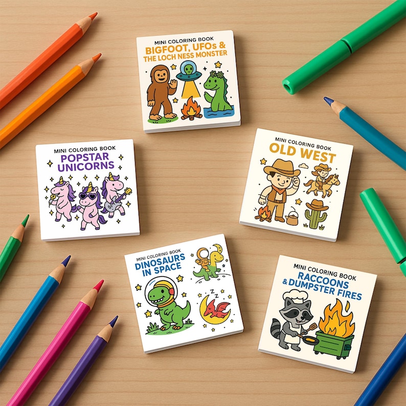May include: A collection of mini coloring books with various themes, including unicorns, Bigfoot, dinosaurs, and raccoons. The books are surrounded by colorful pencils and markers on a wooden surface. Each book features a unique cartoon illustration.