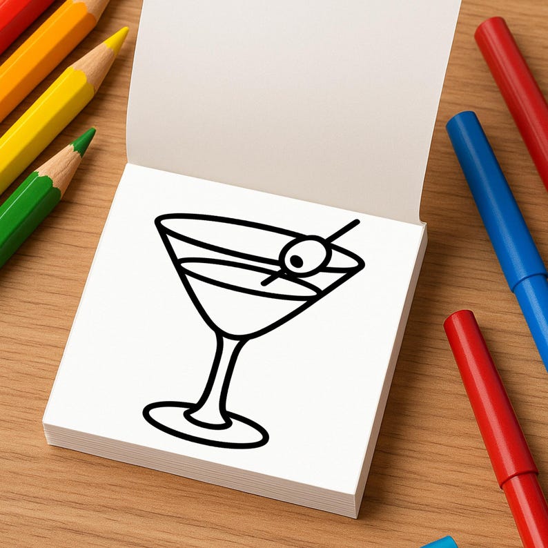 An open mini coloring book displays a simple line drawing of a martini glass with an olive on a toothpick.