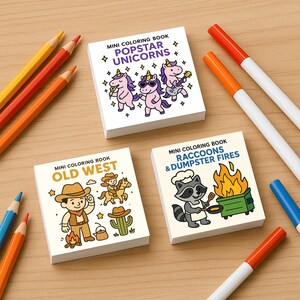 May include: Three mini coloring books with the titles "Popstar Unicorns", "Old West", and "Raccoons & Dumpster Fires" are displayed on a wooden surface. Colored pencils and markers in various colors are scattered around the books. The books feature cartoon illustrations.
