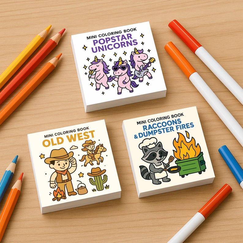 May include: Three mini coloring books with the titles "Popstar Unicorns", "Old West", and "Raccoons & Dumpster Fires" are displayed on a wooden surface. Colored pencils and markers in various colors are scattered around the books. The books feature cartoon illustrations.