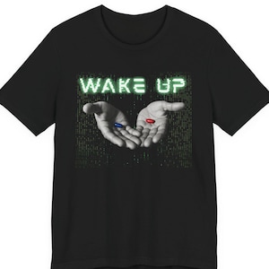 May include: Black t-shirt featuring a graphic of two hands holding a blue and a red pill, with the words "WAKE UP" in green above. The design has a matrix-style background.
