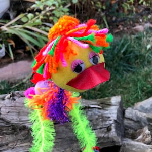 Könnte beinhalten: Eine skurrile, handgefertigte Entendekoration. Die Ente hat einen gelben Körper, Kulleraugen und einen roten Schnabel. Ihr Kopf ist mit orangefarbenem, rotem und rosa Garn geschmückt, und sie hat grüne, lila und rosa Garnbeine. Die Ente hängt vor einem natürlichen Hintergrund.