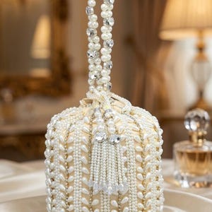 Può includere: Una borsa cilindrica color crema caratterizzata da file verticali di perle bianche a goccia e cristalli trasparenti. La borsa presenta un manico di perle, una chiusura a coulisse con nappa di perle e una base circolare strutturata.