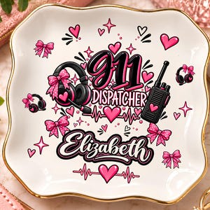 Puede incluir: Un plato decorativo cuadrado de cerámica blanca con borde dorado que presenta un estampado gráfico con el texto 911 DISPATCHER y Elizabeth. El diseño incluye auriculares negros, un walkie-talkie, lazos rosas y corazones rosas con un latido.