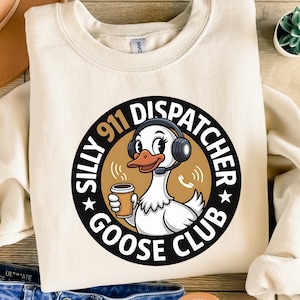Può includere: Felpa color crema con grafica circolare. La grafica presenta un'oca dei cartoni animati con cuffie e una tazza di caffè. Il testo "Silly 911 Dispatcher Goose Club" è in nero.