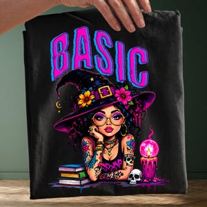 Puede incluir: Una camiseta negra con el gráfico de una persona con sombrero de bruja, gafas redondas y tatuajes. El texto BASIC está impreso en letras grandes rosas y azules arriba. El diseño incluye una pila de libros, una calavera y una vela rosa brillante.