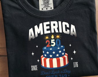 May include: Black t-shirt with the word "AMERICA" above a two-tiered cake decorated with stars and stripes, and the number "250". The shirt also includes the text "SINCE 1776" and "Don't Look a Day * OVER 249*".
