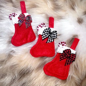 May include: Three small red felt Christmas stockings with white lace cuffs and hanging loops. Each stocking features a miniature candy cane tucked into the cuff and a decorative fabric bow in either plaid or gingham patterns attached to the front.