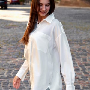 May include: A white shirt with a collar, chest pocket, and long sleeves. The shirt is worn with light blue jeans. The person is walking on a cobbled street.