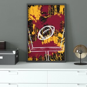 May include: Framed art print featuring a football and goalpost design. The background has a splatter effect in yellow, burgundy, and black. The football and goalpost are outlined in white.