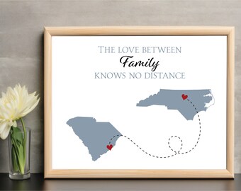 May include: A framed print with a white background and the text "The love between family knows no distance". The print features a map of North Carolina and South Carolina with a dotted line connecting the two states. There are red hearts on the map to indicate the location of the two states.