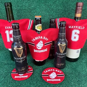 Könnte beinhalten: Mehrere Guinness Stout-Flaschen sind mit roten, Trikot-förmigen Flaschenhüllen der Tampa Bay Buccaneers geschmückt. Die Flaschen stehen auf roten Untersetzern mit dem Buccaneers-Logo. Ein Trikot zeigt "EVANS" und "13", ein anderes "MAYFIELD" und "6".