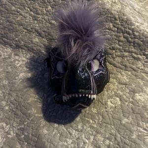May include: A black and purple decorative mask with a feathered mohawk. The mask has large purple eyes, white teeth, and a textured surface. The mask is resting on a gray floral patterned surface.