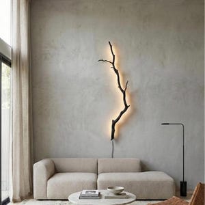 May include: A black, tree branch-shaped wall light with a warm, yellow backlight. The light is mounted on a light grey wall. The room features a neutral-toned sofa, a round coffee table, and a black floor lamp, creating a modern, minimalist aesthetic.