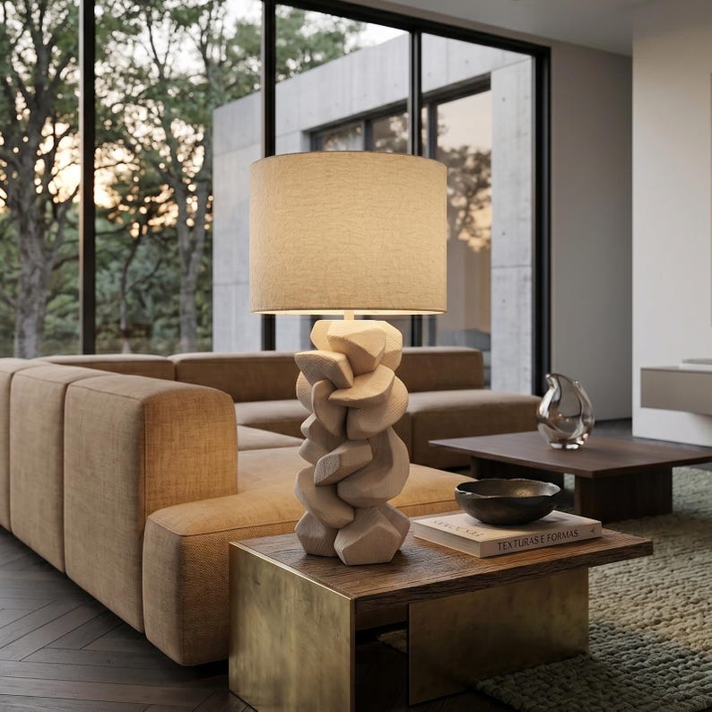 May include: A beige fabric lampshade sits atop a light-colored, abstract wooden lamp base. The lamp is on a wooden coffee table with a brass base, next to a brown sofa. The room has large windows and a modern design.