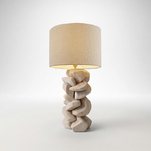 May include: A modern table lamp with a beige fabric shade. The base is made of light-colored wood, carved in an abstract, stacked design. The lamp is lit, casting a warm glow.