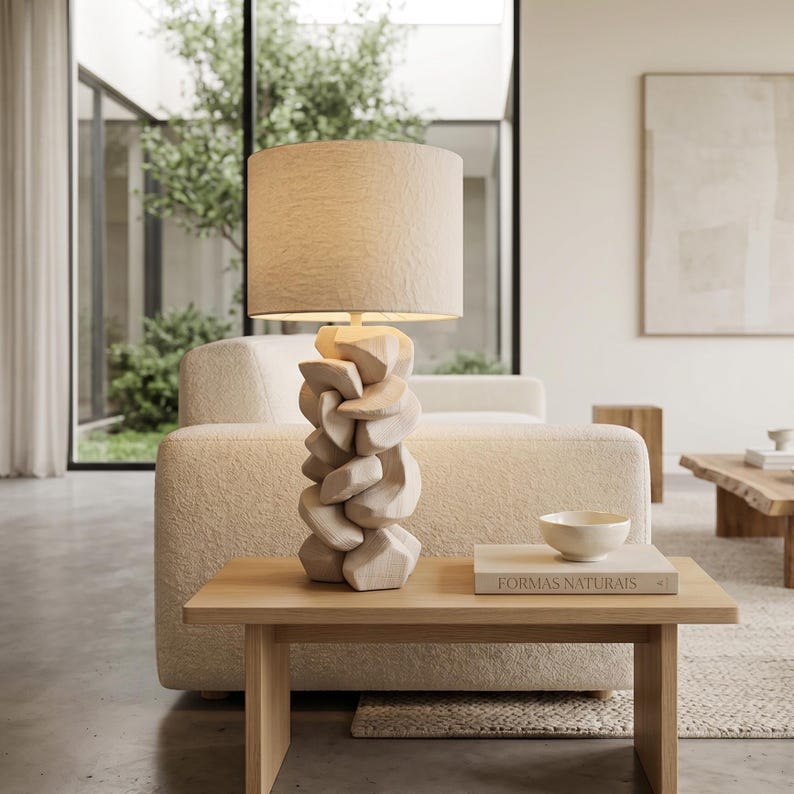 May include: A beige table lamp with a light-colored fabric shade. The lamp base is made of carved wood in a stacked, geometric design. The lamp sits on a light wood table with a book and a small bowl.
