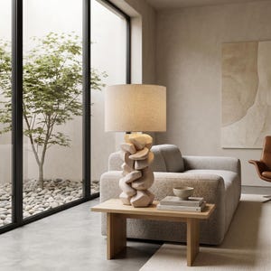 May include: A modern table lamp with a beige linen shade and a unique, carved wooden base. The lamp sits on a light wood side table, next to a sofa. The room has a neutral color palette and a minimalist design.