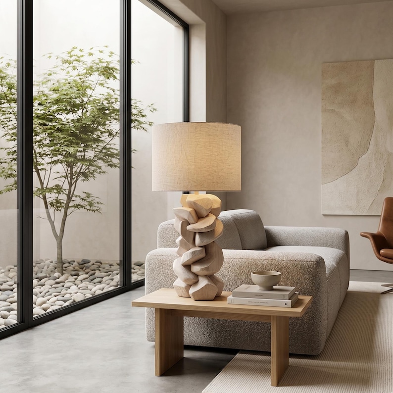 May include: A modern table lamp with a beige linen shade and a unique, carved wooden base. The lamp sits on a light wood side table, next to a sofa. The room has a neutral color palette and a minimalist design.