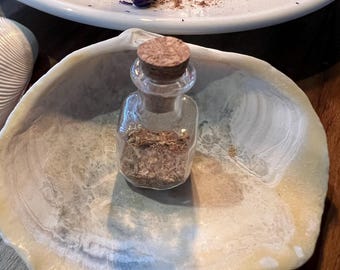 May include: A small, clear glass bottle with a cork stopper containing a light brown granular substance, resting inside a large, off-white, textured seashell. The shell has a smooth, pearlescent interior and a natural, irregular outer edge.