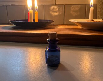 May include: A small, square-shaped cobalt blue glass bottle with a cork stopper. The bottle has a narrow neck and a wider base, containing a dark, granular substance inside. The glass is translucent, reflecting light from nearby candles.