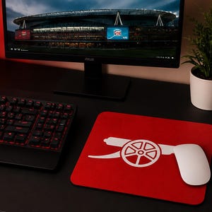 May include: A rectangular red mouse pad featuring a white Arsenal Football Club cannon logo. The mouse pad is positioned on a black desk next to a black computer keyboard and a white computer mouse, with a monitor displaying a stadium in the background.