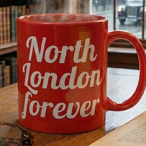 Può includere: Una tazza di ceramica rossa con la scritta bianca "North London forever". La tazza è piena di una bevanda calda, e il vapore sale dalla parte superiore. La tazza è su una superficie di legno.