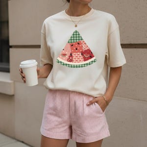 May include: Cream-colored short-sleeved t-shirt featuring a watermelon slice design with a green gingham rind and patchwork details in shades of red and pink. The model is wearing pink and white striped shorts.