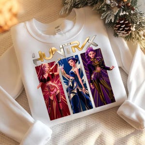 Peut inclure: Sweat-shirt blanc avec un motif de trois personnages féminins stylisés et le texte doré "HUNTRIX". Les personnages portent des tenues rouges, bleues et violettes. Le sweat-shirt a des manches longues et un col rond.