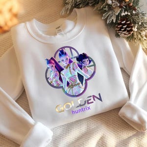 Può includere: Felpa bianca con una grafica di tre figure femminili stilizzate con capelli viola e blu, inserite in un design floreale. La parola "GOLDEN" è stampata in oro, con "huntrix" sotto in viola.