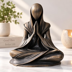 May include: A bronze-colored sculpture of a faceless figure in a hooded robe, seated in a cross-legged position with hands pressed together in a prayer gesture. The figure features a smooth, rounded head and draped fabric textures throughout the body.