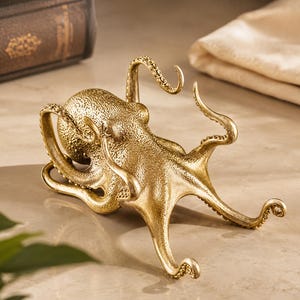 May include: A gold-toned metal octopus figurine featuring a textured mantle and eight curved, detailed tentacles. The sculpture displays a polished metallic finish with visible suction cup patterns along the underside of each extended arm.