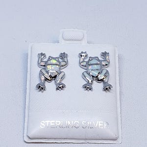 Pode incluir: Um par de brincos de sapo em prata esterlina. Cada brinco apresenta um design de sapo com um centro semelhante a opala. Os brincos são exibidos em um cartão branco com o texto "STERLING SILVER".