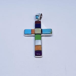 May include: A silver cross pendant with inlaid rectangular stones in various colors, including turquoise, orange, green, and purple. The cross has a bail at the top for a chain. The pendant is likely made of sterling silver.