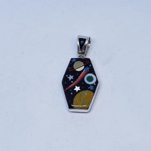 May include: A coffin-shaped pendant featuring a black inlay with a space-themed mosaic design. The inlay includes a gold-colored planet with rings, a green and white circular planet, a yellow planet, and several small white stars and dots.