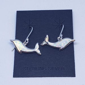 May include: A pair of sterling silver dolphin earrings. Each earring has a dolphin shape with a white and iridescent opal-like inlay. The earrings hang from silver fish hook style ear wires. The words "STERLING SILVER" are printed on the black card.