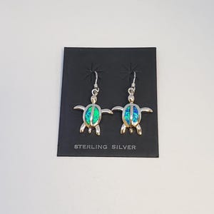 May include: A pair of sterling silver dangle earrings shaped like sea turtles. Each turtle features a vibrant blue and green iridescent inlay on its shell. The earrings are attached to a black display card with the text STERLING SILVER printed.