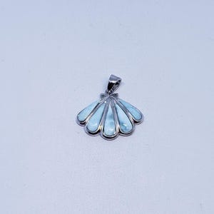 May include: A silver pendant in the shape of a seashell, featuring light blue larimar stones. The pendant has a bail for a chain and is set against a white background. The design is reminiscent of ocean themes.