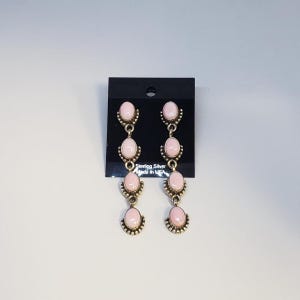 May include: A pair of sterling silver dangle earrings featuring four oval-shaped pink stones each, arranged in a vertical column. The stones are set in silver bezels with beaded edges. The earrings are displayed on a black card labeled Sterling Silver Made in USA.