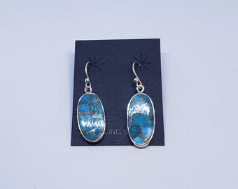 May include: A pair of sterling silver earrings with oval-shaped turquoise stones. The stones have a blue and gold matrix pattern. The earrings are displayed on a black card.
