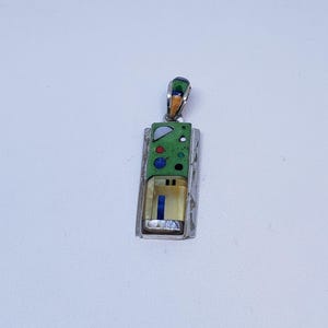 May include: A rectangular pendant with a silver frame and a green, textured center. The pendant features colorful inlaid designs, including blue, red, and white accents. The top has a small loop for a chain. The pendant is approximately 1.5 inches long.