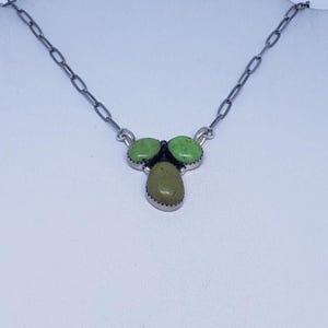 May include: A silver-toned necklace featuring three green stone pendants. Two round stones are positioned above a single, oval-shaped stone. The necklace chain is a series of interconnected links, creating a simple yet elegant design.