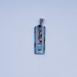 Peut inclure: Pendentif rectangulaire avec bélière et cadre argentés. Le pendentif présente un fond turquoise avec des sections incrustées de pierres rouges, blanches et noires. Le design est géométrique et symétrique, avec une finition polie.