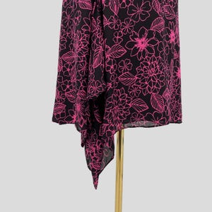 May include: Black and pink floral print fabric. The fabric features a repeating pattern of pink flowers and leaves on a black background. The fabric is draped over a gold-colored stand, showcasing its asymmetrical hemline.