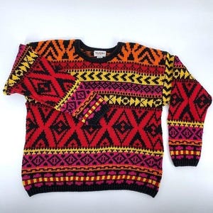 May include: A colorful, patterned sweater in red, black, yellow, orange, and pink. The sweater features a geometric design with diamond and zigzag patterns. The cuffs and neckline are black. The sweater is made of a soft, knitted material.