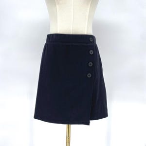 May include: A dark navy blue skirt with a wrap-style design. The skirt features three dark buttons on the side and a straight hemline. The skirt is displayed on a mannequin.