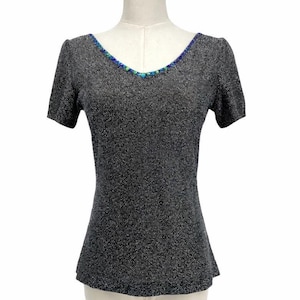 Può includere: Un top grigio a maniche corte con scollo a V. Lo scollo è impreziosito da perline blu e verdi. Il top ha un aspetto strutturato e scintillante ed è esposto su un manichino.