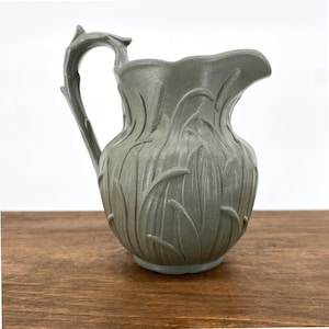 Puede incluir: Una jarra de cerámica gris con un asa decorativa y un diseño floral. La jarra tiene una abertura ancha y mide aproximadamente 18 cm de altura.