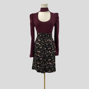 May include: A burgundy long-sleeve dress with a high neckline and a keyhole cutout. The dress features a black pleated skirt with a paisley pattern in beige and white. The dress is displayed on a mannequin.