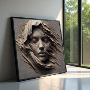 Könnte beinhalten: Ein gerahmtes Kunstwerk mit einem skulptierten Gesicht mit geschlossenen Augen, das in Stoff gehüllt ist. Das Kunstwerk ist in Beige- und Grautönen gehalten und hat ein strukturiertes Aussehen. Der Rahmen ist schwarz, und das Kunstwerk lehnt an einer weißen Wand in der Nähe eines Fensters.